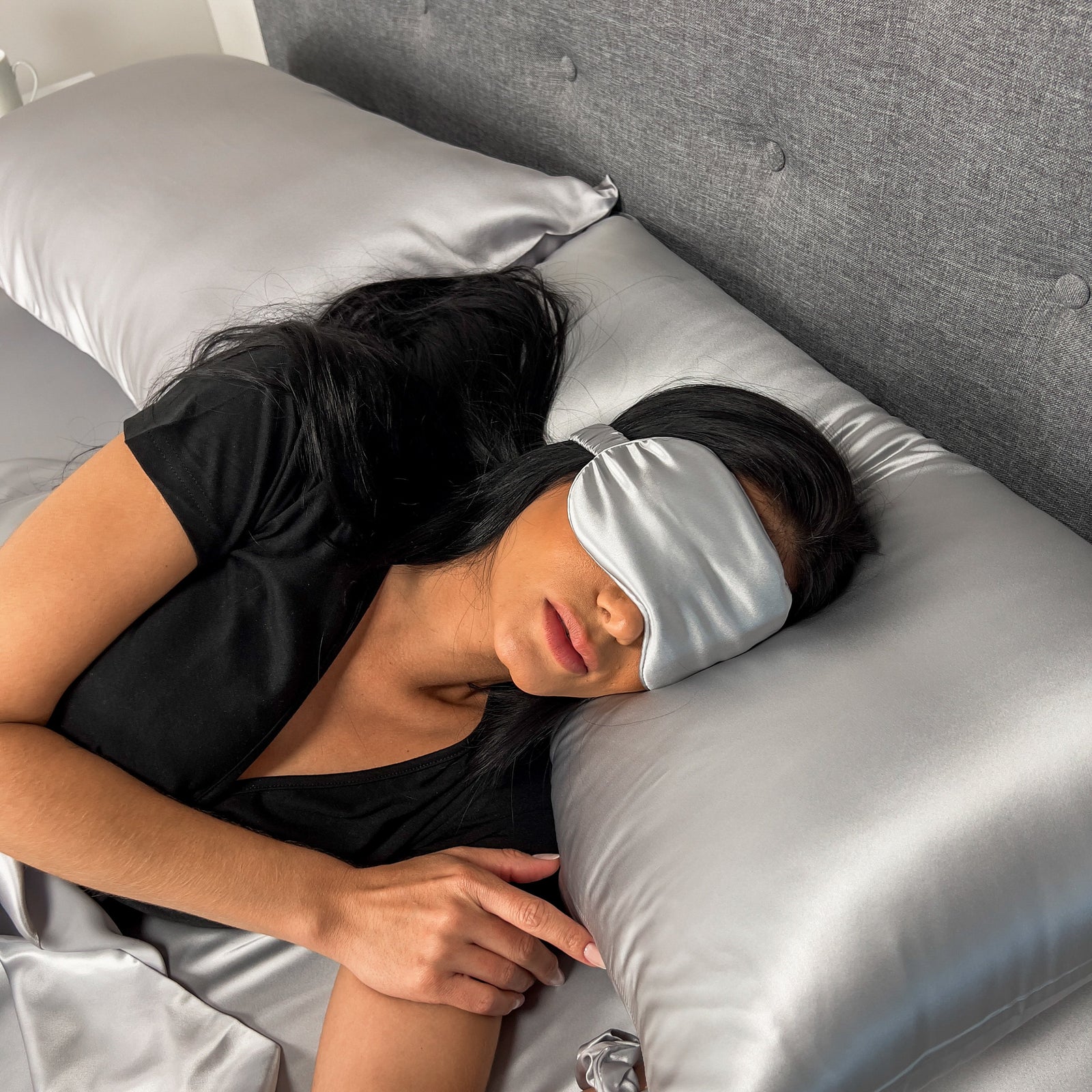 Woman resting on a silver silk pillowcase with a matching sleep mask, lying on the bed.