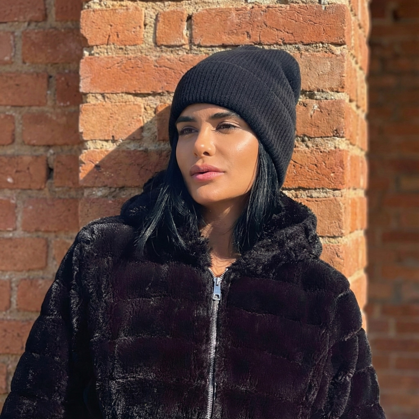 Woman wearing a black coat and beanie against a brick wall