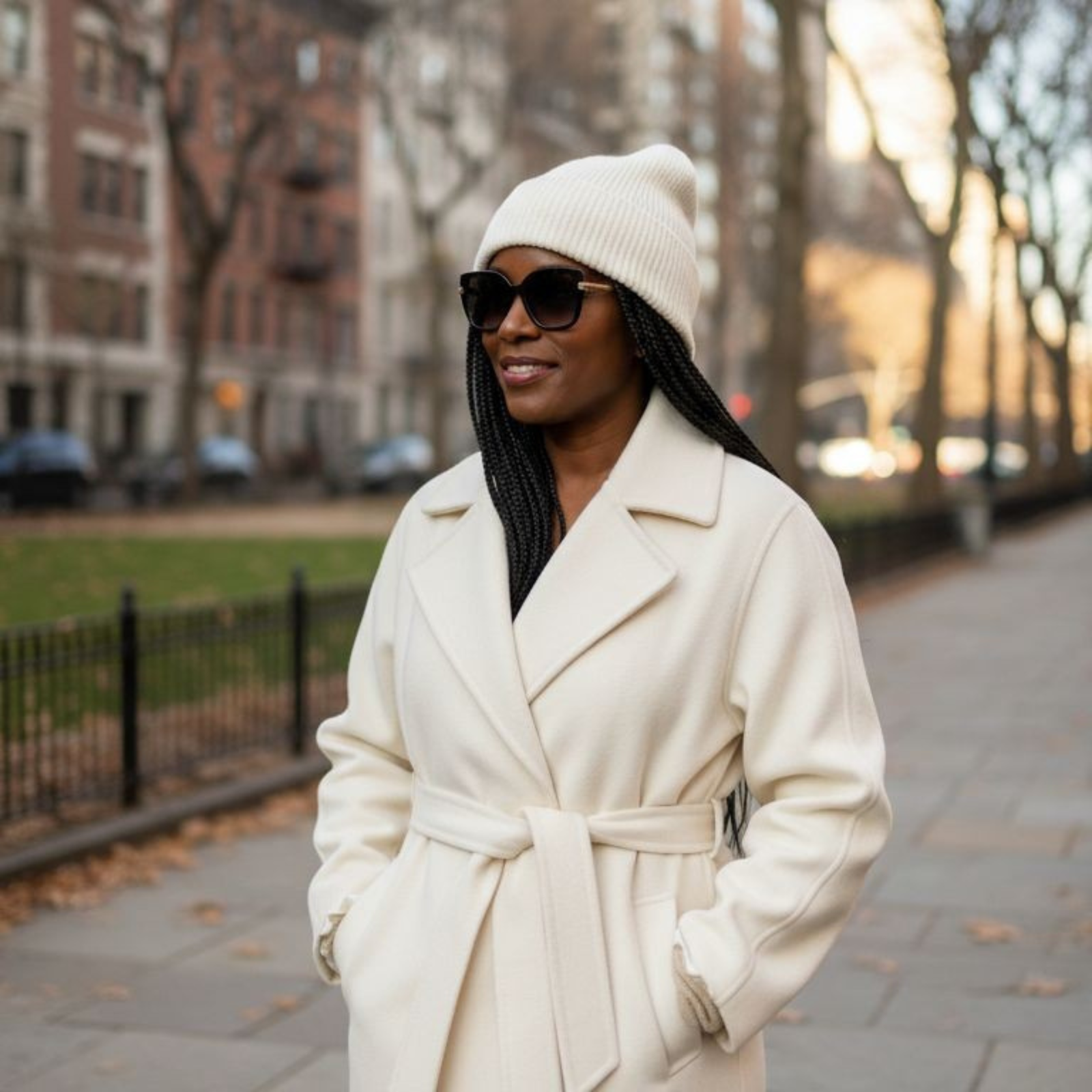 Woman in an ivory coat and beanie standing on a city street.